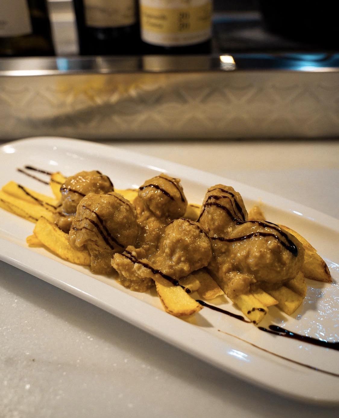 Ración en salsa servida sobre patatas en plato alargado, con las botellas de la barra al fondo